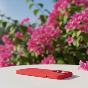 May include: A red phone case with a sleek design, featuring a camera cutout and button details. The case is set on a white surface, with vibrant pink flowers and green foliage in the background, under a clear blue sky.