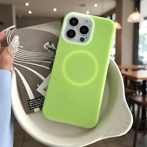Puede incluir: Una funda de teléfono verde brillante con un anillo de cámara blanco y un diseño circular en la parte posterior. La funda está colocada en un plato blanco curvado. Una tarjeta con texto y un dibujo es parcialmente visible.