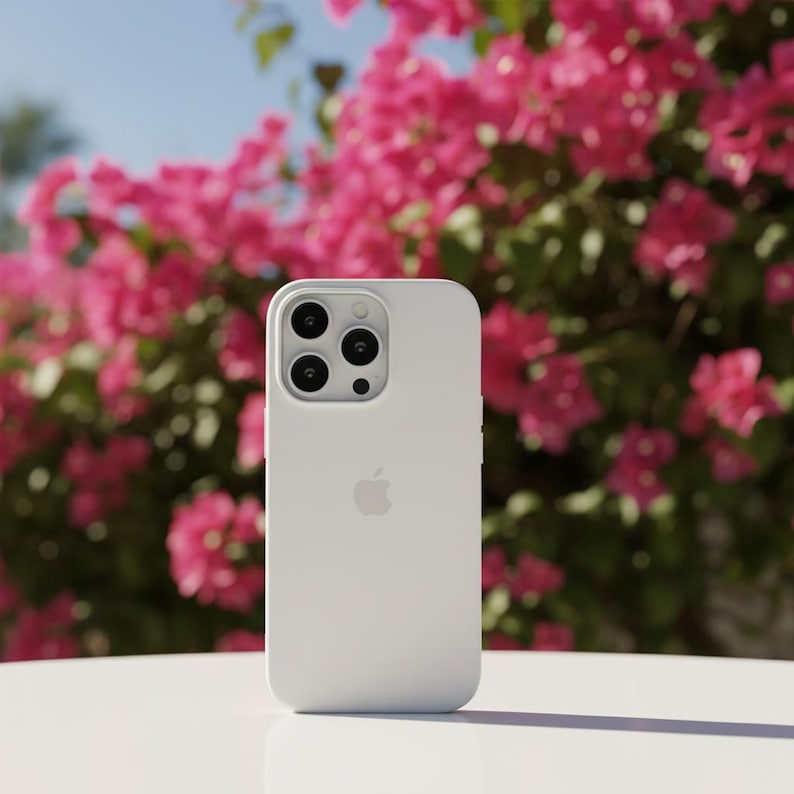 May include: A white smartphone case with a matte finish. The case has a camera module with three lenses and a flash. The Apple logo is visible on the back. The background features pink flowers and a blue sky.