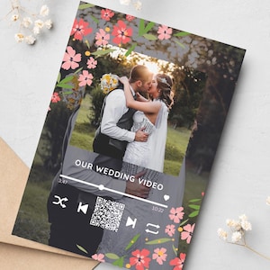 Può includere: Una carta rettangolare con una fotografia centrale di una coppia che si bacia in un campo, incorniciata da un bordo di fiori rosa e foglie verdi. La carta mostra un'interfaccia di lettore musicale digitale con il testo OUR WEDDING VIDEO e un codice QR.