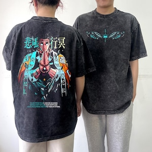 May include: Two dark gray t-shirts with graphic designs. One shirt features a colorful anime-style illustration on the back with Japanese text and the quote "WE ARE PROUD TO LIVE AND DIE AS HUMAN BEINGS..." The other shirt has a teal and blue design.