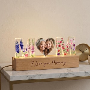 Könnte beinhalten: Ein dekoratives beleuchtetes Schild mit der Aufschrift "MOM", floralen Designs und einem herzförmigen Fotorahmen. Die Holzbasis trägt die Aufschrift "I Love you Mommy". Die Buchstaben sind mit warmweißen Lichtern beleuchtet.
