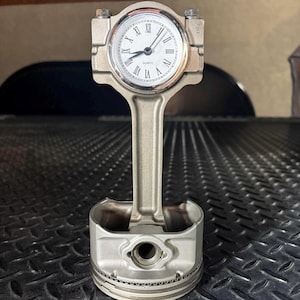 Puede incluir: Un reloj de escritorio construido a partir de un pistón de motor metálico y una biela. La esfera del reloj es circular con fondo blanco, números romanos negros y manecillas negras, con la palabra QUARTZ impresa en letras negras pequeñas.