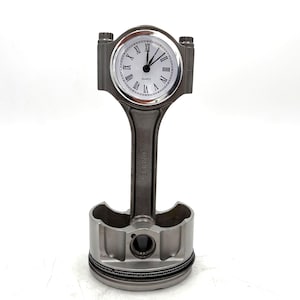 May include: A unique clock made from a repurposed engine piston. The clock face is white with black Roman numerals and hands. The piston is silver-colored and stands upright, making it a distinctive desk or shelf decor item.