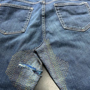 May include: Close-up of the back of blue denim jeans with visible wear and tear. The jeans have been repaired with a visible mending technique using yellow, orange, and red thread. The repair is on the left knee.