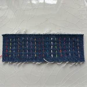 May include: A rectangular piece of denim fabric with colorful vertical stitching. The stitches are in various colors, including red, yellow, green, blue, and purple. The edges of the fabric are frayed, with loose threads visible.