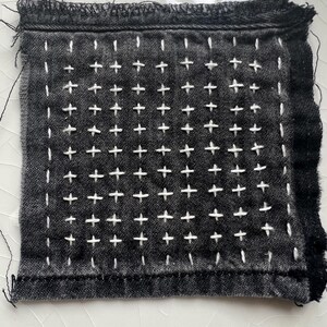 May include: A square, dark gray fabric patch with visible white stitching in a grid pattern. The stitching forms small crosses across the surface. The edges of the fabric are frayed, with loose threads.