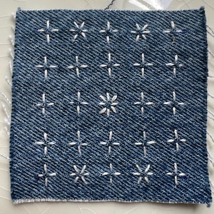 May include: Square denim fabric with a grid pattern of white cross-stitch embroidery. The blue denim has a textured appearance, and the white thread creates a contrasting design. The edges of the fabric are unfinished, with loose threads.