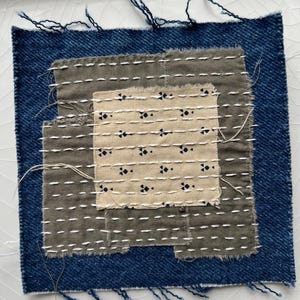 May include: A square textile piece featuring a patchwork design. The base is a dark blue denim, overlaid with gray and beige fabric squares. White stitching and decorative black floral patterns add detail. Frayed edges give a rustic, handcrafted appearance.