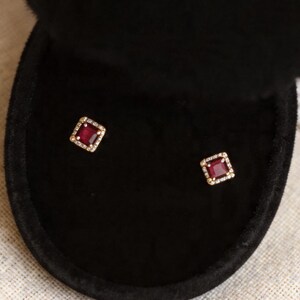 May include: A pair of square-cut ruby earrings displayed in a black velvet jewelry box. The earrings have a gold-colored border with small, sparkling accents. A hand holds the open box, with a pearl necklace in the background.