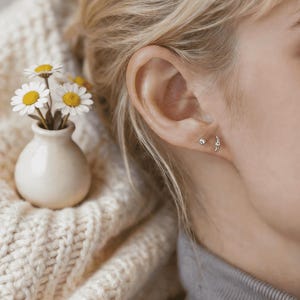 Pode incluir: Um close-up de uma orelha com dois pequenos brincos de prata. Um é um pino simples, e o outro tem um design curvo. Um pequeno vaso branco com margaridas está à esquerda, ao lado de um suéter de malha creme.