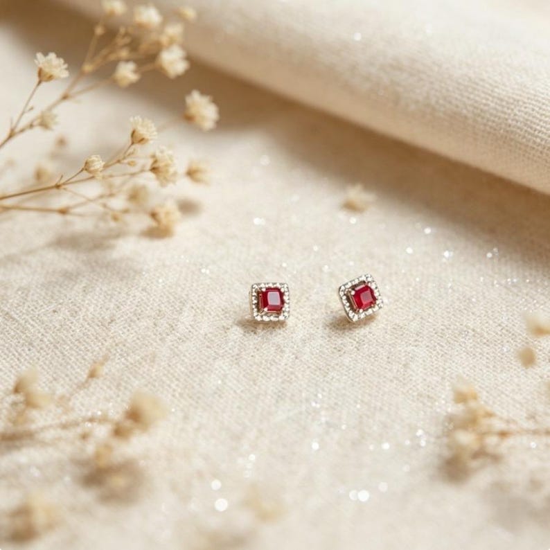 Puede incluir: Un par de pendientes cuadrados con una piedra central roja vibrante, rodeada por un halo de piedras m&aacute;s peque&ntilde;as y claras. Los pendientes est&aacute;n sobre una superficie clara y texturizada, con delicados acentos florales.
