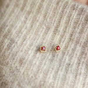 Puede incluir: Un par de pendientes peque&ntilde;os y cuadrados. Cada pendiente presenta una gema roja vibrante engastada en un marco dorado. Los pendientes se muestran sobre una superficie beige claro texturizada.
