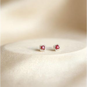 Puede incluir: Un par de pendientes cuadrados con una piedra central roja vibrante, rodeada por un halo de piedras m&aacute;s peque&ntilde;as y claras. Los pendientes est&aacute;n engarzados en un metal plateado y descansan sobre una superficie suave de color crema. El dise&ntilde;o es simple pero elegante.