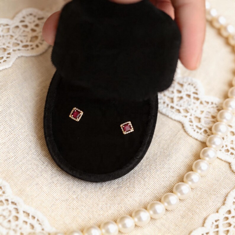 May include: A pair of square-cut ruby earrings displayed in a black velvet jewelry box. The earrings have a gold-colored border with small, sparkling accents. A hand holds the open box, with a pearl necklace in the background.