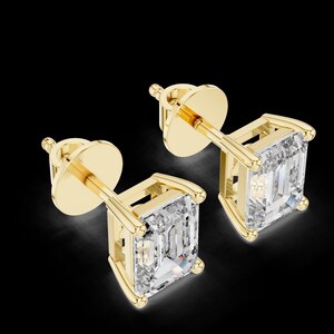 Könnte beinhalten: Ein Paar goldene Ohrstecker mit quadratisch geschliffenen, klaren Edelsteinen. Die Ohrringe haben ein klassisches Design mit Stecker und Verschluss. Die Edelsteine sind in einer goldenen Lünettenfassung gefasst, vor schwarzem Hintergrund.