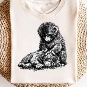 Puede incluir: Camiseta color crema con una ilustración detallada en blanco y negro de dos monos. Uno está sentado y acicalando al otro, que está acostado. El diseño está centrado en la camiseta.
