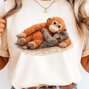 Puede incluir: Camiseta color crema con un gráfico de dos monos abrazándose. El mono más grande es naranja con una cara sonriente, y el mono más pequeño es gris. El diseño está impreso en una camiseta suave e informal.