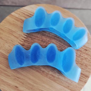 Puede incluir: Dos separadores de dedos de los pies de color azul claro hechos de un material flexible. Cada separador tiene cinco secciones redondeadas diseñadas para encajar entre los dedos. Los separadores están sobre una superficie redonda de madera.