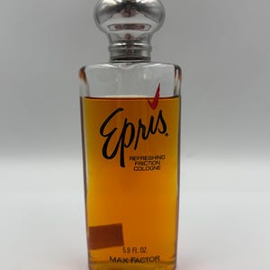 May include: A clear glass bottle of Epris Refreshing Friction Cologne by Max Factor. The bottle contains a golden-amber liquid and has a silver-colored, spherical cap. The label features black script and red accents. The bottle holds 5.9 fl oz.