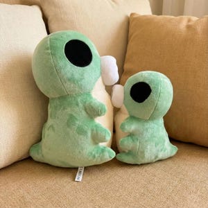 May include: Two plush alien toys in light green with large black eyes and small arms and legs. The toys are sitting on a beige couch with beige pillows in the background. The toys have a white accent on the side.