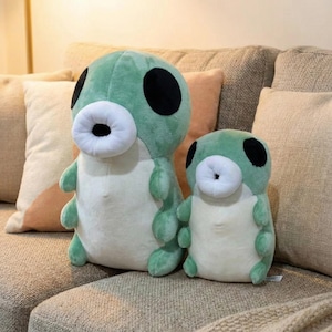 May include: Two plush toys shaped like green creatures with large black eyes and white mouth-like features. The toys are in different sizes and are sitting on a beige couch with pillows.
