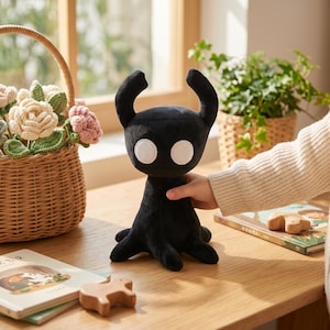 Pode incluir: Um brinquedo de pelúcia preto com olhos brancos e chifres curvos. O brinquedo tem um corpo arredondado e quatro pernas pequenas. A pelúcia é segurada por uma mão. Uma cesta de vime com flores de crochê e um livro estão ao fundo.
