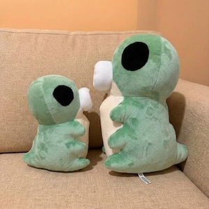 May include: Three plush alien toys in varying sizes. The aliens are light green with large black eyes and small white appendages. They are sitting on a beige couch.
