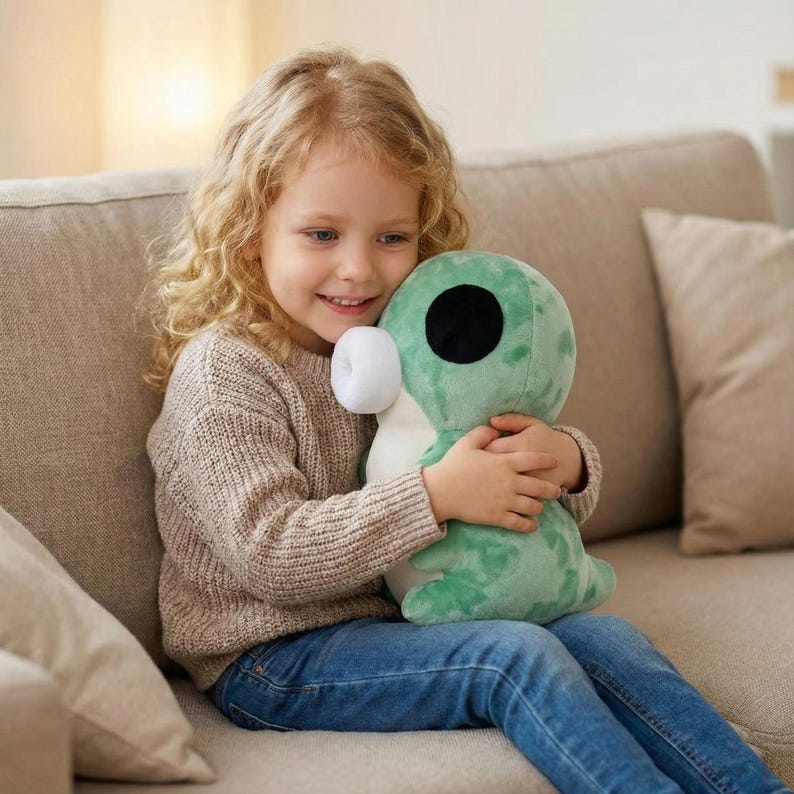 May include: A young child wearing a beige sweater and blue jeans embraces a green plush toy with a large black eye and white accents. The child is sitting on a beige sofa, smiling.