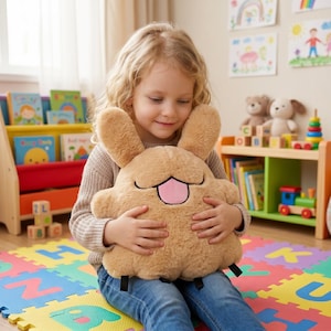 Pode incluir: Um peluche de cor castanha clara com orelhas compridas e uma língua cor-de-rosa de fora. O brinquedo tem uma textura macia e felpuda e uma forma arredondada e compacta. Apresenta linhas pretas bordadas simples para os olhos e a boca.