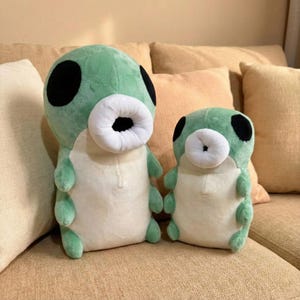 May include: Two plush caterpillar toys. The larger one is light green with black eyes and a white mouth. The smaller one has the same design. Both are sitting on a tan couch with beige pillows.
