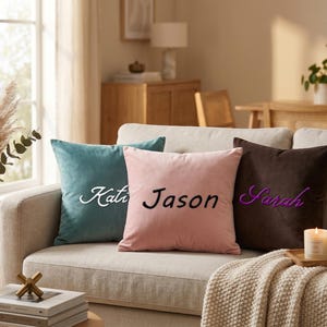 Op de afbeelding: Drie gepersonaliseerde fluwelen sierkussens in teal, roze en bruin, elk geborduurd met een naam: "Kate", "Jason" en "Sarah". De kussens liggen op een lichtgekleurde bank, met een kaars en een mok op een houten dienblad.