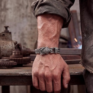 May include: A silver-toned bracelet with an ornate design featuring a central decorative element and a clasp. The bracelet is worn on a wrist, showcasing its intricate details and craftsmanship. The background suggests a workshop setting.