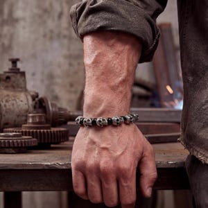 Könnte beinhalten: Ein Armband mit silbernen Totenkopfperlen und schwarzen Perlen. Das Armband wird an einem Handgelenk getragen, wobei die Hand auf einer Metalloberfläche ruht. Der Hintergrund umfasst Industriemaschinen und eine Person, die ein braunes Hemd trägt.