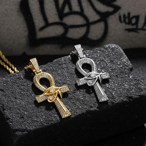 May include: Two Ankh pendants, one gold-toned and one silver-toned, each encrusted with small, sparkling crystals. The gold pendant features an eye design. Both are displayed on a dark, textured surface, with a graffiti-covered background.