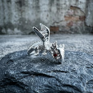Könnte beinhalten: Ein silberner Drachenring mit detaillierten Merkmalen und einem roten Edelsteinauge. Der Drache hat ausgebreitete Flügel und ist auf einer dunklen, strukturierten Oberfläche platziert. Ein Accessoire im Fantasy-Stil.