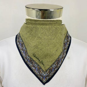 Könnte beinhalten: Ein salbeigrünes Halstuch mit einem detaillierten schwarzen Paisley-Muster. Das Tuch hat einen dekorativen Rand mit blauen, braunen und weißen Paisley-Motiven sowie einen dunkelblauen Saum. Der Schriftzug Christian Dior ist in Schwarz gedruckt.