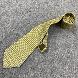 Puede incluir: Una corbata amarilla con un patrón tejido en forma de cesta. La corbata está enrollada en la parte superior, mostrando el patrón. La corbata tiene una etiqueta marrón en la parte posterior. La corbata está sobre una superficie gris.