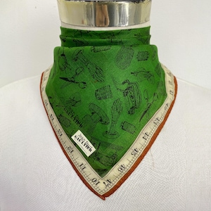 May include: Green square scarf with a repeating black pattern of sewing tools and hands. The scarf has a white border with a brown edge and measurement markings. The label reads "Jean Paul Gaultier."