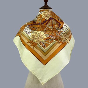 May include: A square scarf featuring a cream-colored border and a central pattern of orange, brown, and purple floral motifs. The design includes geometric shapes, leopard print accents, and detailed flower illustrations arranged in a symmetrical layout.