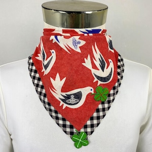 Peut inclure: Un bandana triangulaire à fond rouge orné d'illustrations d'oiseaux blancs, chacun portant un petit logo orbe bleu. Le bandana présente une bordure à carreaux vichy noirs et blancs et deux appliqués en tissu en forme de trèfle vert.