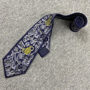 May include: Navy blue silk tie with a repeating pattern of musical instruments, teapots, and other decorative elements in gold and gray. The tie is rolled up at the end, showing the pattern. The tie is laying on a gray surface.