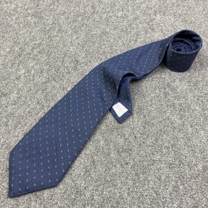 May include: A navy blue tie with a repeating white pattern of small, stylized shapes. The tie is lying on a gray surface, with the end rolled up. The tie has a classic pointed end and a white label.