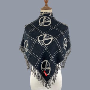 May include: A black triangular shawl featuring a white grid pattern and repeating white circular spiderweb motifs. The edges are finished with long, thin black and white fringe, and a small red rectangular tag is attached to the lower corner.