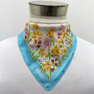 Peut inclure: Un foulard carré à imprimé floral avec une bordure bleu clair et le nom de la marque "KENZO" imprimé dans le coin. Le foulard présente un fond blanc avec des fleurs jaunes, roses et violettes.
