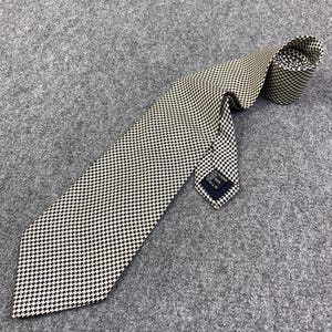 Può includere: Una cravatta a fantasia pied-de-poule bianca e nera. La cravatta è realizzata in tessuto e arrotolata nella parte superiore. Una piccola etichetta è visibile vicino alla punta della cravatta. La cravatta è appoggiata su una superficie grigia.