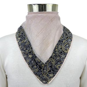 May include: A Christian Dior neck scarf featuring a solid light pink centre with subtle diagonal lines and a wide black border decorated with a detailed gold and white paisley pattern. The scarf is folded into a triangular shape around the neck.