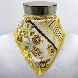 Op de afbeelding: Een sjaal met gele, witte en bruine patronen, met bloemen- en geometrische ontwerpen. De sjaal heeft een gele rand met witte stippen en de tekst "PIERRE BALMAIN". De sjaal wordt op een mannequin getoond.