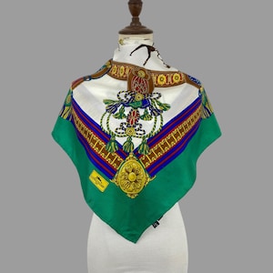 Puede incluir: Un pañuelo de seda cuadrado con un centro blanco y un motivo central ornamentado de borlas y cuerdas doradas, azules y verdes. El borde es de un verde vibrante con un patrón geométrico dorado. Una etiqueta de Longchamp es visible en la parte inferior izquierda.