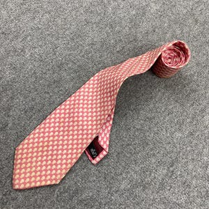 Puede incluir: Una corbata rosa con un patrón blanco repetido. La corbata está enrollada en el extremo yace sobre una superficie gris. La corbata está hecha de un material suave y tiene un diseño clásico.